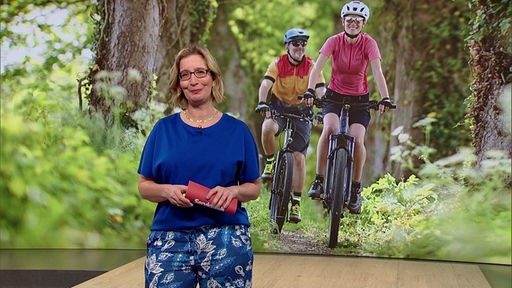 Das Bild zeigt Yvonne Willicks im Servicezeit-Studio. Im Hintergrund sind zwei Fahrradfahrer:innen abgebildet.